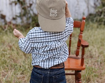 May include: A tan baseball cap with the text big bro embroidered in white on the front. The cap features a curved brim and metal eyelets. It is worn with a white and black plaid long-sleeve button-down shirt and dark blue denim jeans.
