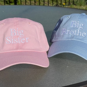 May include: Two baseball caps, one pink and one light blue, with white embroidered text. The pink cap says "Big Sister" and the blue cap says "Big Brother".