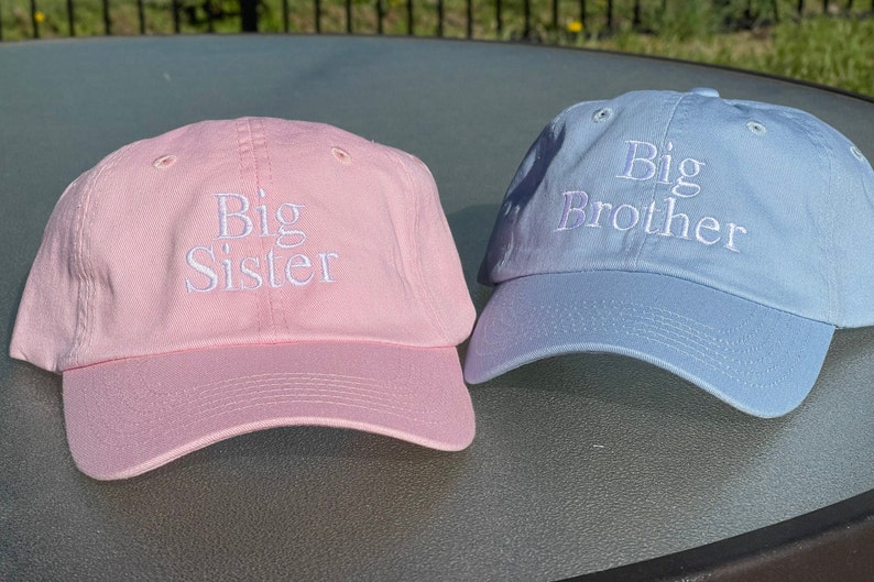May include: Two baseball caps, one pink and one light blue, with white embroidered text. The pink cap says "Big Sister" and the blue cap says "Big Brother".