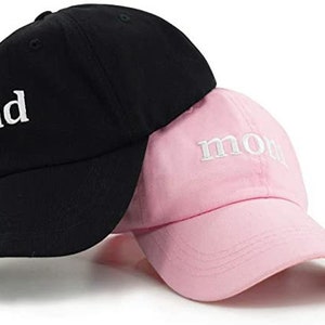 Puede incluir: Dos gorras de b&eacute;isbol, una negra con la palabra "dad" bordada en blanco, y una rosa con la palabra "mom" bordada en blanco.