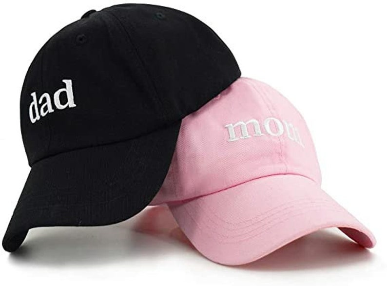 Puede incluir: Dos gorras de b&eacute;isbol, una negra con la palabra "dad" bordada en blanco, y una rosa con la palabra "mom" bordada en blanco.