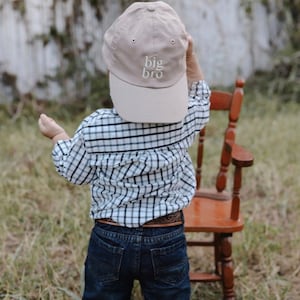 May include: A young child wearing a beige baseball cap with the text "big bro" embroidered on the front. The child is wearing a plaid shirt and blue jeans.
