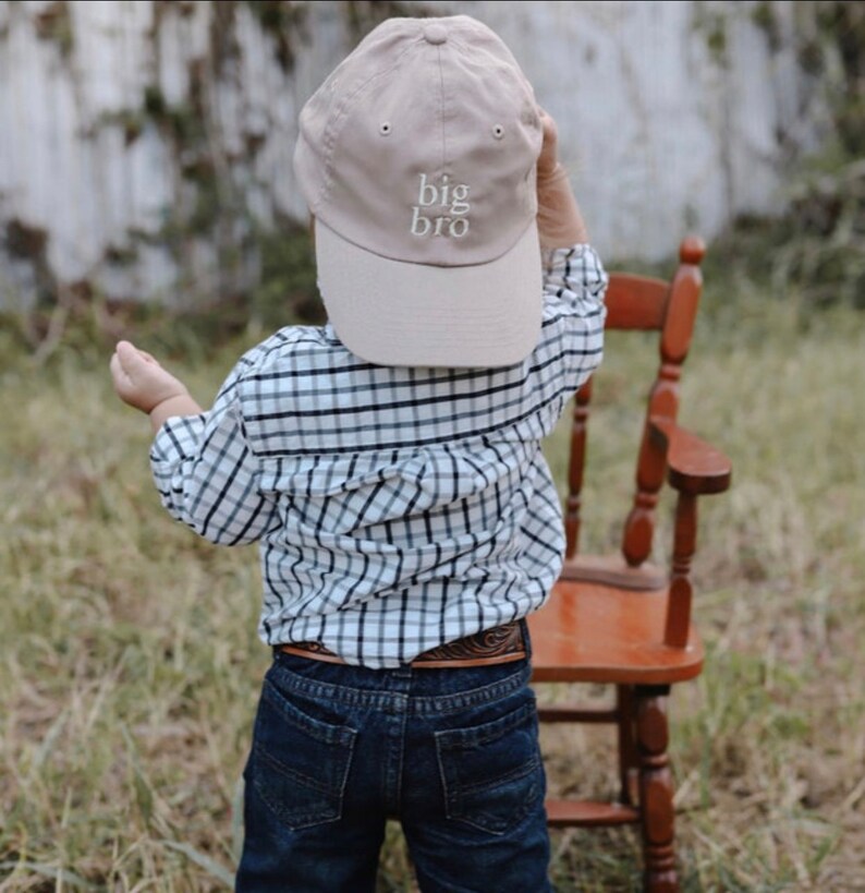 May include: A young child wearing a beige baseball cap with the text "big bro" embroidered on the front. The child is wearing a plaid shirt and blue jeans.