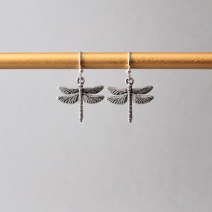 May include: Two silver dragonfly earrings. Each earring has a detailed dragonfly pendant with outstretched wings and a slender body. The earrings hang from simple silver hooks, showcasing a nature-inspired design.