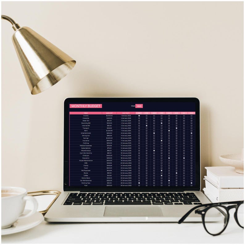 May include: A laptop computer with a black screen displaying a monthly budget spreadsheet. The spreadsheet includes categories for income and expenses, with columns for the amount, date, and whether the expense has been paid. The laptop is on a white desk with a gold lamp, a cup of coffee, and a stack of books.
