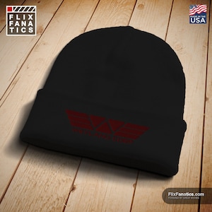May include: Black knit beanie with a red graphic of a stylized wing design and the text "WEYLAND CORP". The hat is made in the USA and is displayed on a wooden surface. The FlixFanatics logo is in the upper left corner.