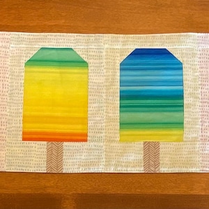May include: A table runner with six vibrant popsicle designs. Each popsicle displays a different gradient of colours, such as blue, pink, yellow, and green. The popsicles are set against a cream-coloured background with a textured pattern.