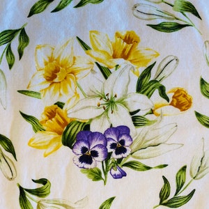 May include: A white fabric with a floral print featuring yellow daffodils, white lilies, purple pansies, and green leaves.