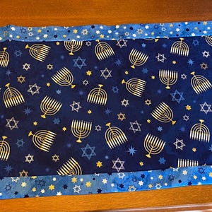 Puede incluir: Camino de mesa de Janucá azul y dorado. El centro presenta una tela azul oscuro con menorás y estrellas doradas. El borde es azul claro con patrones de estrellas doradas y blancas. El camino de mesa es rectangular con extremos angulados.