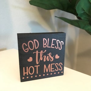 May include: A black wooden sign with the text "God Bless This Hot Mess" in rose gold lettering. The sign has a distressed look and is decorated with small hearts and dots.