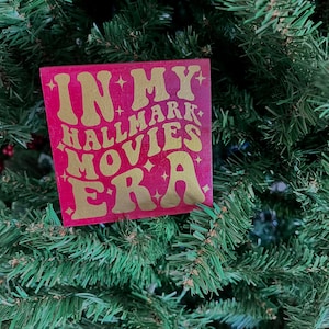May include: A red wooden sign with gold lettering that reads "In My Hallmark Movies Era". The sign is hanging on a green Christmas tree.