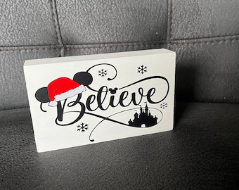 Może przedstawiać: Biały drewniany znak z napisem "Believe" w czarnym, ozdobnym piśmie. Na znaku znajduje się czarna sylwetka Myszki Miki w czapce Świętego Mikołaja i czarna sylwetka zamku Kopciuszka.