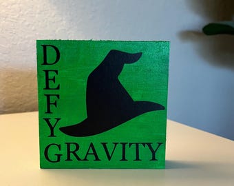 Może przedstawiać: Zielony drewniany blok z napisem "DEFY GRAVITY" czarnymi literami. Na środku bloku znajduje się czarna sylwetka czarodziejskiego kapelusza.
