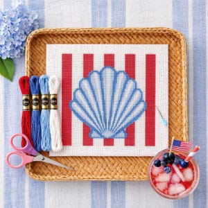 May include: A cross-stitch project featuring a blue and white seashell design on a red and white striped background. The craft supplies include embroidery floss in red, blue, and white, pink scissors, and a cocktail with blueberries and an American flag.