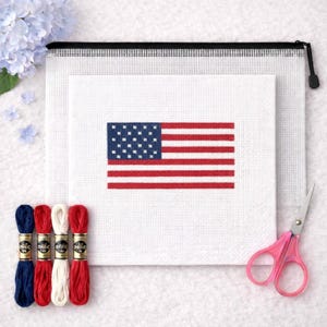 May include: A cross-stitch kit depicting the American flag. The kit includes a white canvas with the flag design, red, white, and blue embroidery floss, pink scissors, and a white mesh zipper pouch. The finished design measures approximately 20 cm by 15 cm.