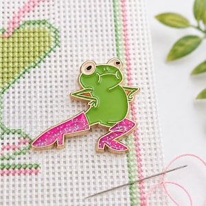 May include: A green frog enamel pin with gold trim and pink glitter boots. The frog is posed with its arms out and legs spread. The pin is on a white canvas with pink and green stitching.