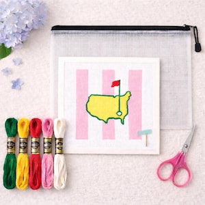 May include: A needlepoint kit featuring a golf course design with a yellow and green map, pink and white stripes, and a red flag. Includes embroidery floss in various colors, a clear zippered pouch, and pink scissors.