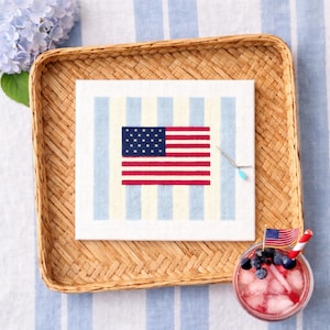 May include: A square needlepoint of the American flag, with red and white stripes and a blue field of stars. The needlepoint is on a striped background and sits in a woven tray. A cocktail with blueberries and a small flag is in the corner.