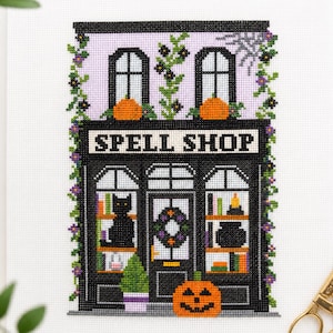 May include: A cross-stitch embroidery on white fabric depicting a storefront labeled SPELL SHOP. The black building features two windows with orange pumpkins, a central door with a wreath, a black cat, a cauldron, and climbing purple flowers.