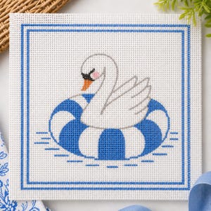 May include: A square needlepoint design featuring a white swan floating on a blue and white striped inner tube. The swan has a gray head, a pink cheek, and an orange beak. The design is framed with a blue border.