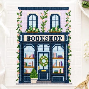May include: Cross-stitch design of a quaint bookshop with a navy blue facade, a lavender-colored upper story, and the word "BOOKSHOP" above the entrance. The windows are adorned with floral accents and the shop's shelves are filled with colorful books.