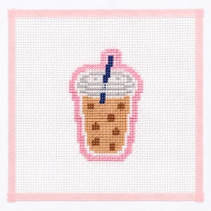 May include: A cross-stitch design of a bubble tea drink in a clear cup with a blue straw. The drink is brown with dark brown boba pearls. The cup is outlined in pink, and the background is white with a pink border.