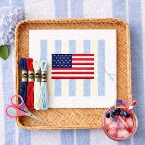 May include: A cross-stitch project featuring the American flag, displayed on a white canvas with blue and yellow stripes. The image includes red, white, and blue embroidery floss, pink scissors, and a refreshing drink with an American flag.