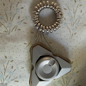 May include: A silver metal coil hair tie and a silver and gold fidget spinner. The fidget spinner has a triangular shape with a central circular component. The hair tie is a spiral of metal.