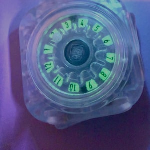 May include: A clear, square-shaped resin object featuring a circular center with a glowing green dial. The dial displays the numbers 3 through 12 in a clockwise arrangement. A small, dark square emblem is positioned in the center of the dial.