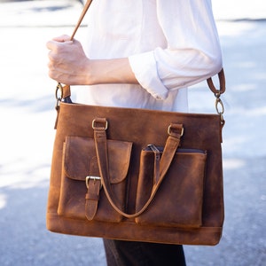 May include: A brown leather satchel bag with a long strap and multiple pockets. The bag has a buckle closure and brass hardware.