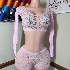 May include: A light pink, open-weave two-piece outfit on a mannequin. The set includes a long-sleeved crop top with a deep V-neck and shorts. The top has a sequined bra-like insert. The shorts are high-waisted.