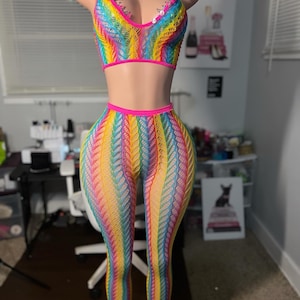 Puede incluir: Un conjunto de dos piezas de crochet a rayas arcoíris con ribete rosa. La parte superior es de estilo halter y los pantalones son largos y ajustados.