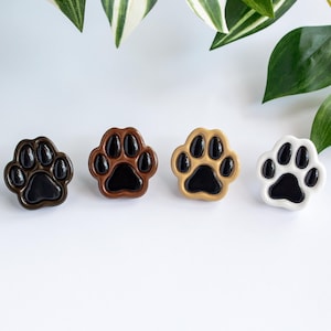 Peut inclure: Quatre boutons de meuble en forme de patte, de couleurs variées : marron foncé, marron moyen, beige et blanc. Chaque bouton a un centre noir, et les boutons sont disposés en rangée. Les boutons sont probablement en céramique ou dans un matériau similaire.