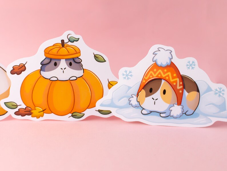 May include: Two illustrated stickers featuring a guinea pig wearing a pumpkin hat and a guinea pig wearing a winter hat. The stickers are on a pink background.