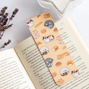 May include: A yellow bookmark with a repeating pattern of cats and bread. The cats are in various poses, including sleeping, sitting, and walking. The bread is in various shapes, including loaves, rolls, and slices. The bookmark is made of a durable material and is perfect for marking your place in a book.