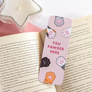 May include: A pink bookmark featuring cat paw prints and the text "YOU PAWSED HERE" in pink. The bookmark is placed in an open book. A star-shaped candle holder is visible.