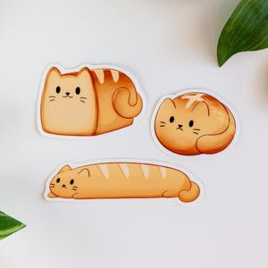 Puede incluir: Tres pegatinas con forma de gato en varias poses. Las pegatinas son en tonos naranja y marrón, que se asemejan al pan. Cada gato tiene una cara linda y simple con ojos y bigotes negros. Las pegatinas están sobre un fondo blanco con hojas verdes.
