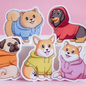 Könnte beinhalten: Fünf Cartoon-Hunde-Aufkleber, die bunte Hoodies tragen. Die Hunde sind ein Mops, ein Corgi, ein Dackel, ein Shiba Inu und ein Zwergspitz.