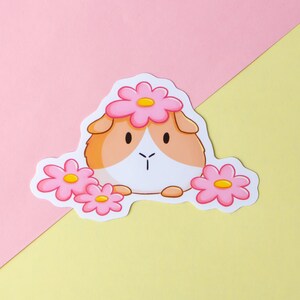 May include: A sticker featuring a cartoon guinea pig wearing a pink flower crown. The guinea pig is brown and white with a pink nose and pink flowers on either side of it.