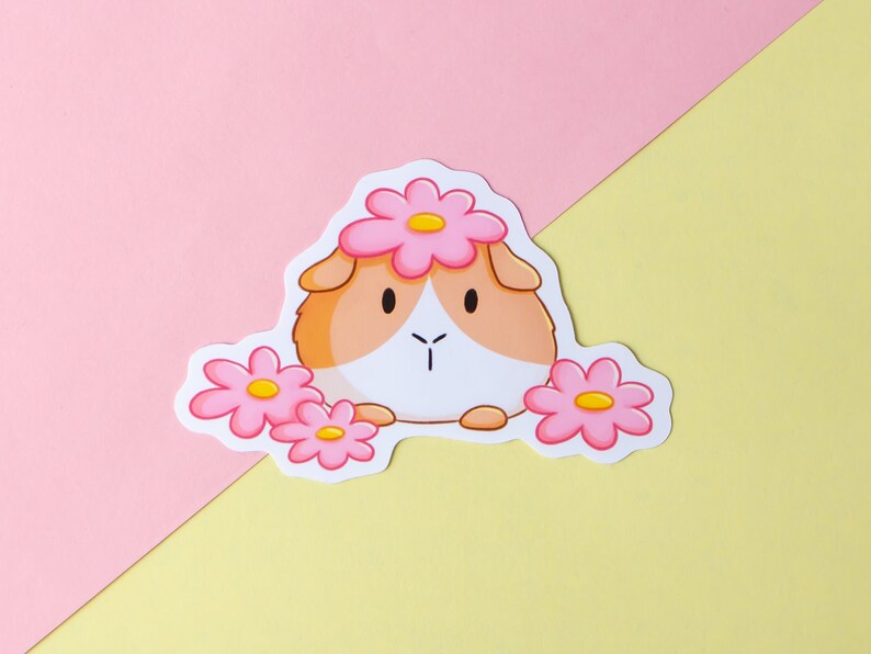 May include: A sticker featuring a cartoon guinea pig wearing a pink flower crown. The guinea pig is brown and white with a pink nose and pink flowers on either side of it.