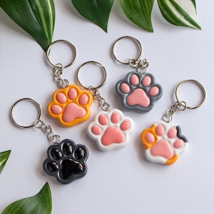 Peut inclure: Plusieurs porte-clés en forme de pattes de chat, de couleurs orange, noir, blanc et gris, chacun avec un coussinet rose. Les porte-clés sont attachés à des anneaux argentés. Le fond est blanc avec des feuilles vertes.