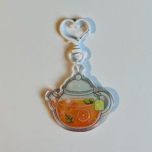 Könnte beinhalten: Ein klarer Acryl-Schlüsselanhänger mit einem Teekannen-Design, gefüllt mit orangener Flüssigkeit, Fruchtscheiben und Minzblättern. Die Teekanne hat einen hellgrünen Deckel und einen kleinen grünen Teebeutel. Der Schlüsselanhänger hat einen weißen herzförmigen Verschluss.