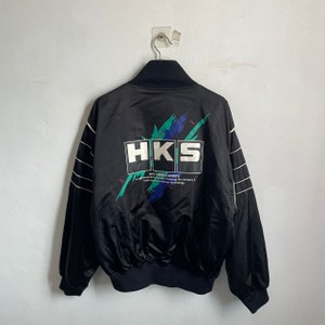 Könnte beinhalten: Schwarze Satin-Bomberjacke mit einem weißen HKS-Logo und einem bunten Grafikdesign auf der Rückseite. Der Text "HKS POWERFUL SPORTS" ist unter dem Logo aufgedruckt.