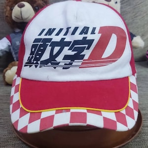 Op de afbeelding: Een witte en rode baseballpet met een geruit patroon op de klep. De voorkant van de pet heeft de tekst "INITIAL D" in zwart en wit met Japanse karakters in zwart.