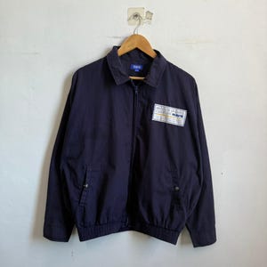 May include: Navy blue zip-up jacket with a collared neckline and two front pockets. The jacket features a white rectangular patch with the text "MOTOR SPORTS CULTURE RAYS". The jacket is hanging on a wooden hanger.