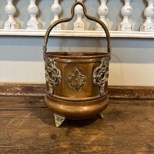 May include: A copper bucket with a brass handle and decorative brass accents. The bucket has a rounded bottom and three feet. The bucket is sitting on a wooden surface.