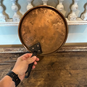 May include: A round copper pan with a black handle. The pan has a textured surface and a dark patina. The handle is attached with rivets. The pan is likely vintage and suitable for cooking.