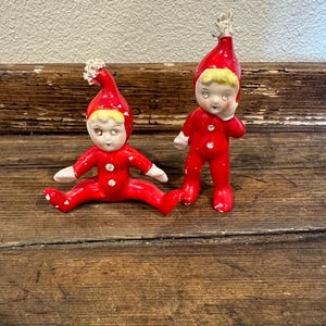 May include: Two red and white ceramic elf figurines. The elf on the left is sitting with its legs crossed and the elf on the right is standing with its right hand on its chin.