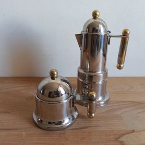 May include: A silver and gold coffee maker and a silver sugar bowl with a lid. Both items have a shiny, polished finish.