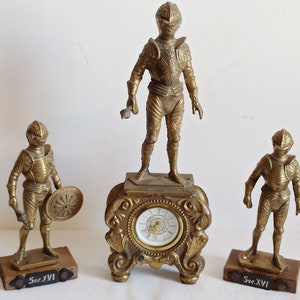 May include: Three gold-toned metal figurines of knights in armour, each standing on a wooden base with the inscription "Sec. XVI". The centre figurine is taller and features a clock face.
